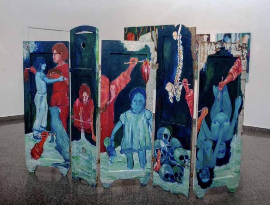 Biombo de cinco folhas de madeira, de alturas diferentes, pintadas em azul, verde e vermelho: figuras azuis e ruivas, um cachorro azul, um coração anatômico, uma coluna vertebral, crânios e braços vermelhos com pincéis, numa sala de paredes brancas.