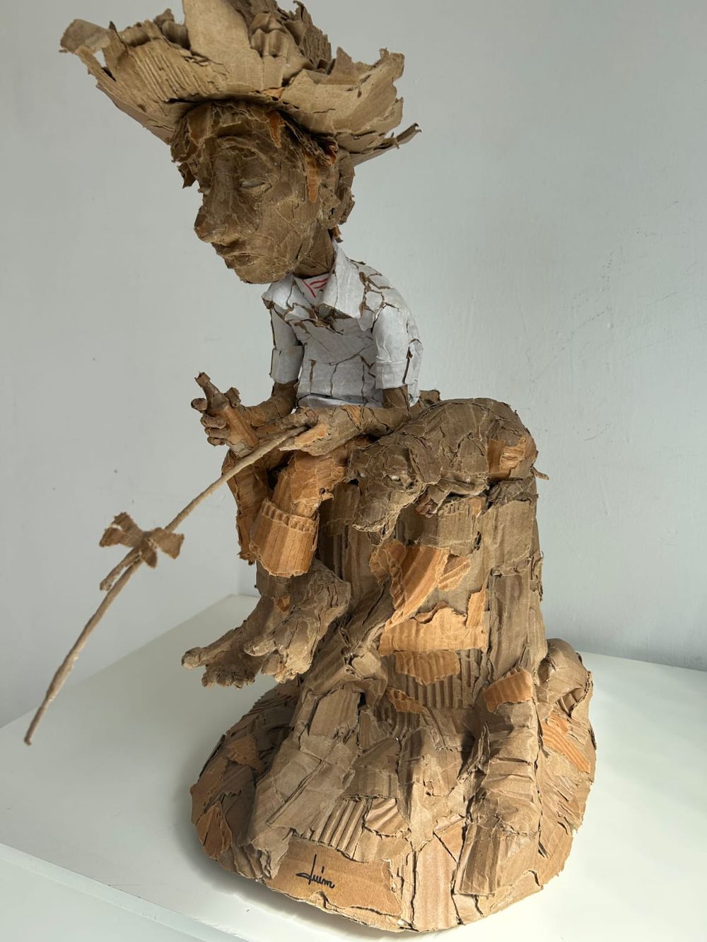 Escultura de papelão de um pescador sentado, de chapéu largo, camisa branca e vara de pesca, com um animalzinho deitado no toco.
